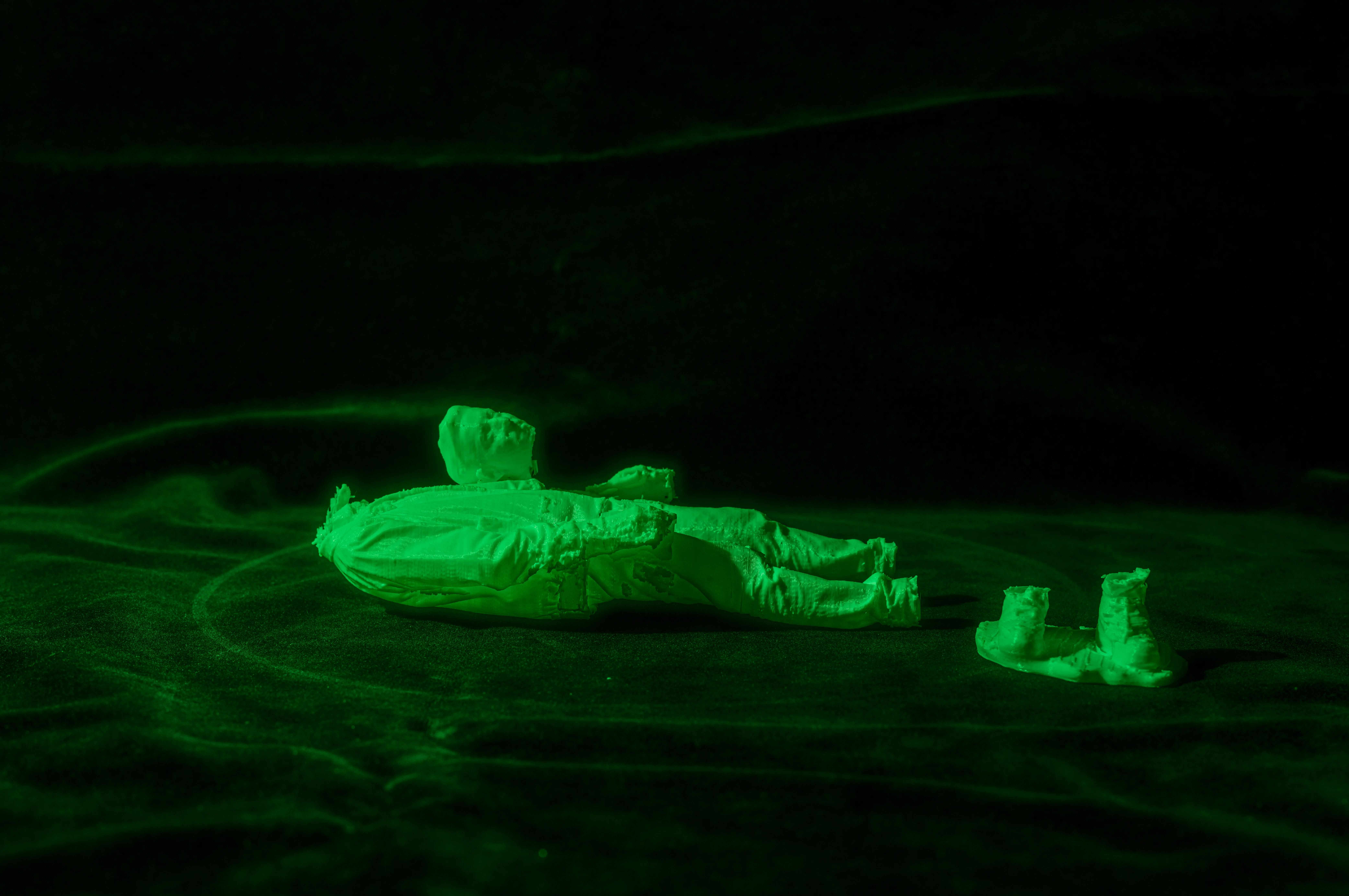 Une photographie une scène sombre éclairée par une lumière verte. Au centre, une figurine humaine semble être allongée sur une surface texturée, rappelant un sol ou un lit recouvert d'un drap. La figurine est cassée la tête et les pieds sont détachés du corps. La lumière verte donne un aspect surnaturel à l'ensemble de la scène, ajoutant une atmosphère mystérieuse et énigmatique.