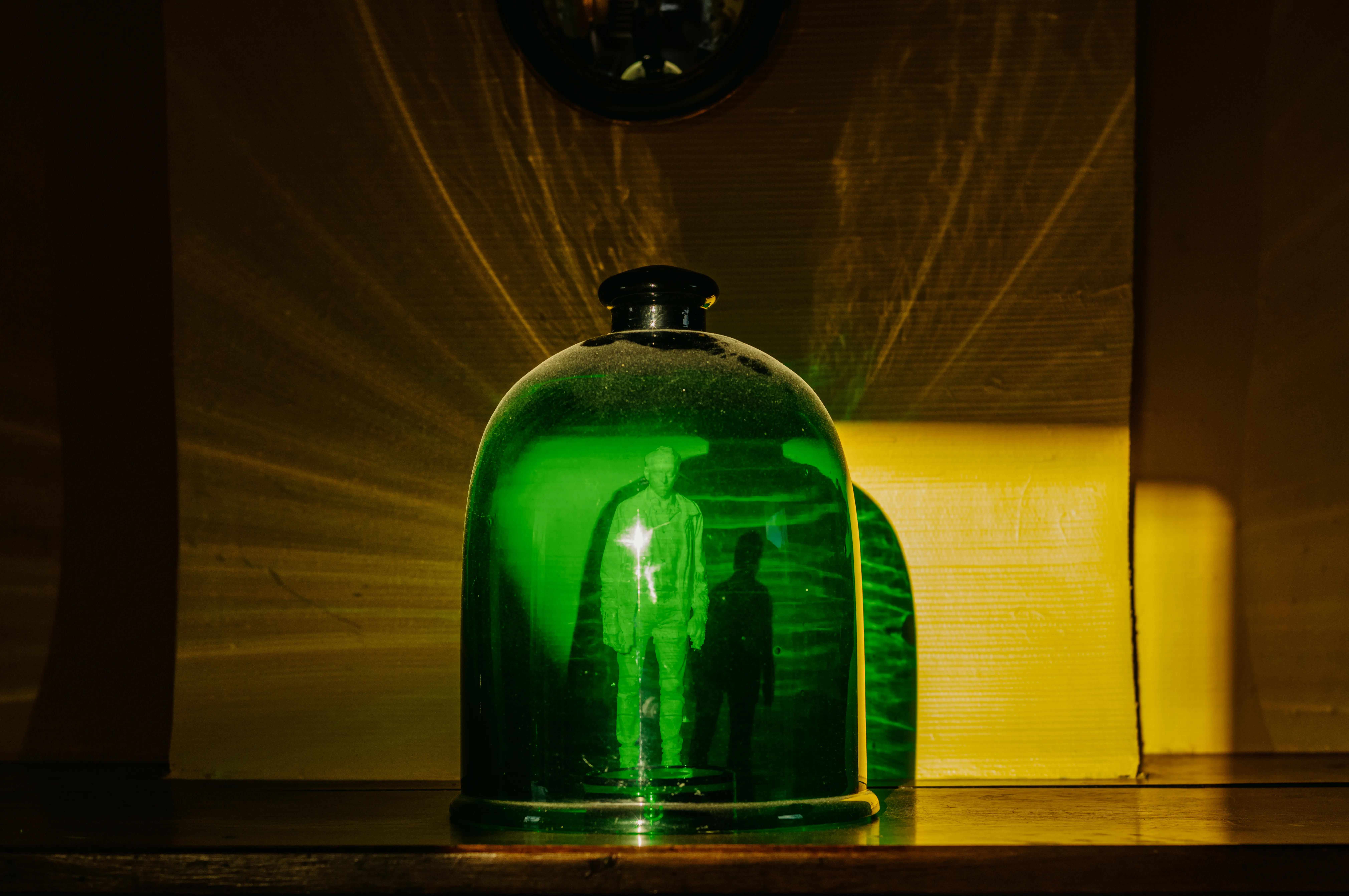 Photographie d'une cloche en verre vert posée sur une surface en bois, éclairée de manière à projeter des ombres dramatiques sur le mur derrière elle. À l'intérieur de la cloche, on distingue une figurine, éclairée d'une lumière qui crée un effet éthéré. L'arrière-plan est partiellement éclairé par une lumière jaune, ajoutant une atmosphère mystérieuse et intrigante à la scène.