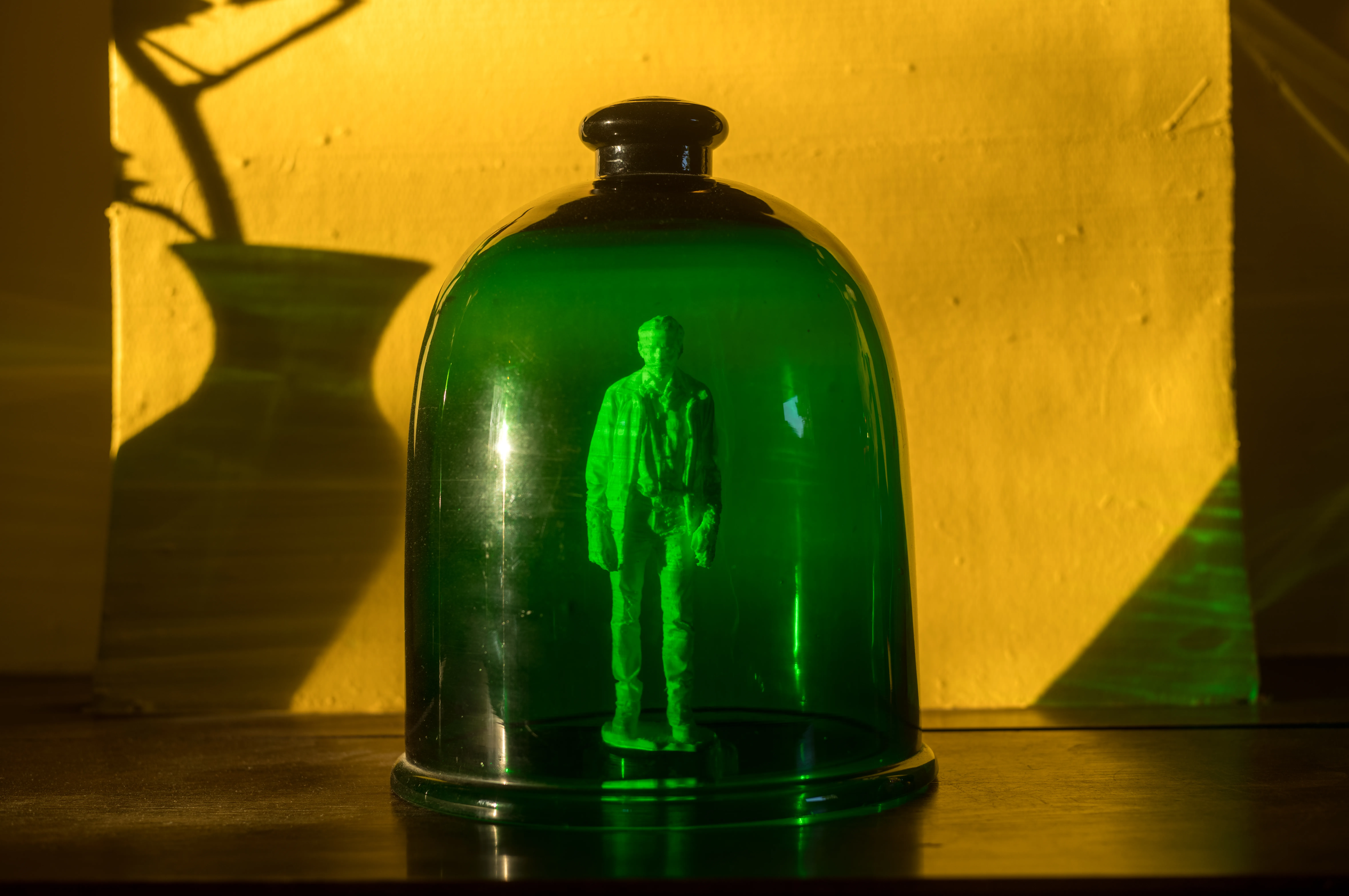 Photographie d'une cloche en verre vert posée sur un support métallique, avec en arrière-plan un papier peint aux motifs floraux colorés. À l'intérieur de la cloche, on distingue la silhouette d'une figure humaine debout, éclairée par une lumière qui traverse le verre, créant un effet mystique. La scène est illuminée par une lumière chaude, renforçant l'ambiance étrange et captivante de l'image.