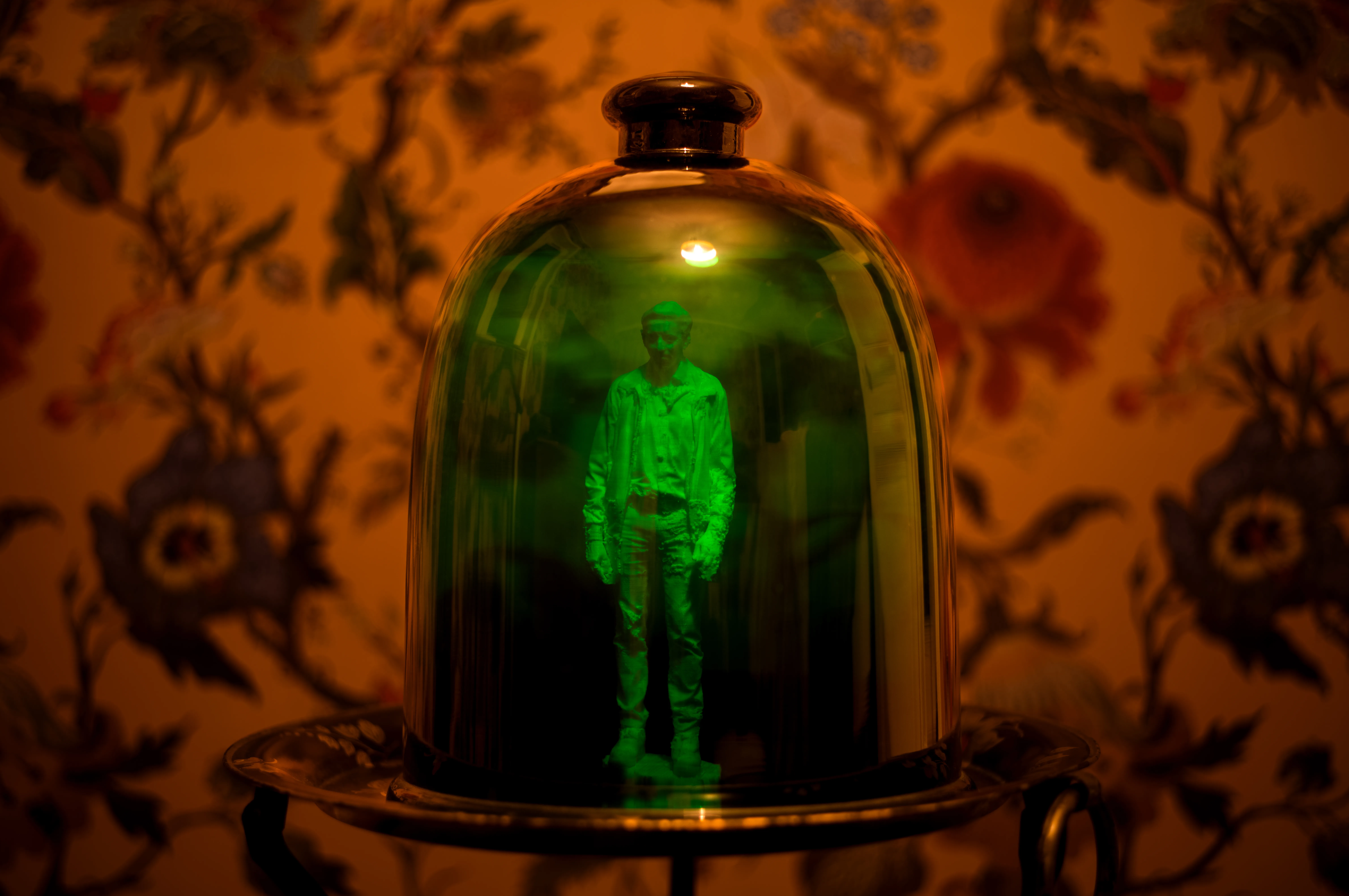 Photographie d'une cloche en verre vert posée sur une surface en bois. À l'intérieur de la cloche, on peut voir figurine debout, éclairée par une lumière qui traverse le verre. L'arrière-plan est d'un jaune lumineux, avec l'ombre d'un objet ressemblant à un vase projetée sur le mur. La combinaison des couleurs et des ombres crée une ambiance mystérieuse et captivante.
